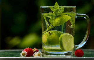 Glas mit aromatisiertem Wasser, darin Limettenscheiben und frische Minze, daneben Erdbeeren und Gänseblümchen auf einem Tisch im Freien
