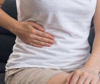 Person in weissem T-Shirt sitzt auf einem dunklen Sofa und legt die Hand auf den rechten Oberbauch, Bereich der Leber